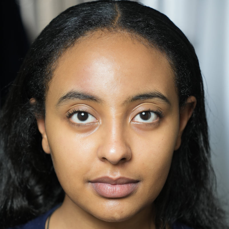A close-up portrait of a woman with dark hair, wearing a navy blue top, set against a soft, out-of-focus gray curtain background after using LED Face Mask.