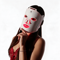 A woman wearing a byTheia Advanced LED light therapy face mask with red glowing light through the eye, nose, and mouth cutouts, set against a bright white background.